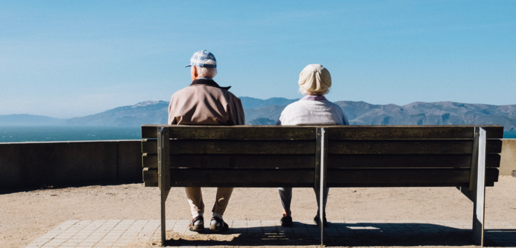 Foto de dos personas mayores, un hombre y una mujer, de espalda a la cámara, sentadas en una banca y mirando el mar. Al fondo se ven cerros y un cielo azul despejado,
