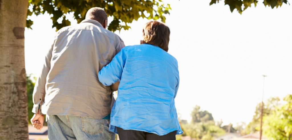 Foto que muestra de espaldas a una mujer de mediana edad ayudando a caminar a un hombre mayor, en un parque.