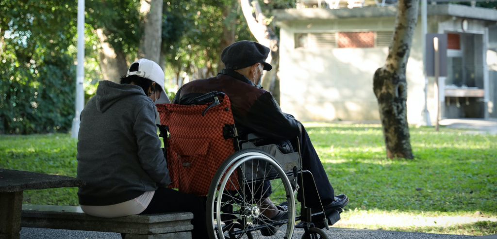Foto de una mujer cuidadora en un parque, sentada en una banca detrás de un hombre mayor en una silla de ruedas. Ambos usan mascarilla.