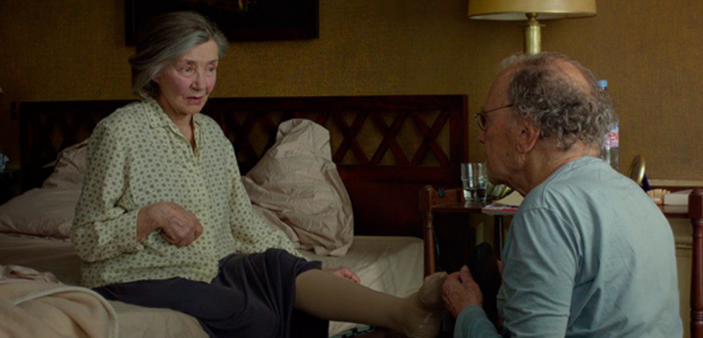 Fotograma de la película Amour que muestra a una mujer mayor en una cama siendo ayudada por un hombre mayor.