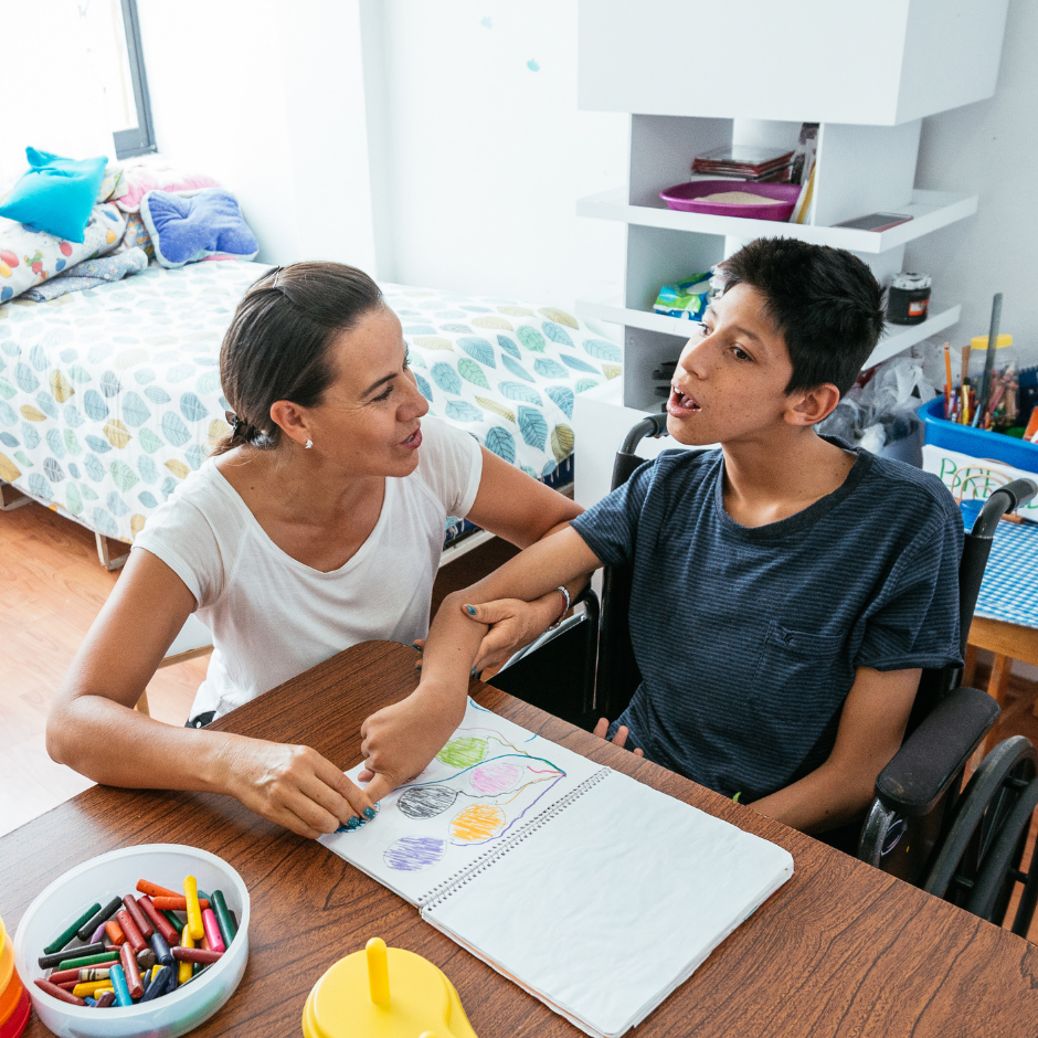 Foto de una madre junto a su hijo con discapacidad intelectual que dibuja en un cuaderno al interior de una habitación de una casa