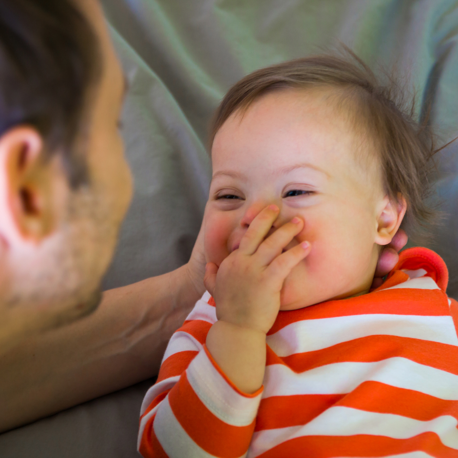 Foto de un bebé con síndrome de down acostado y sonriendo a su papá que lo mira.
