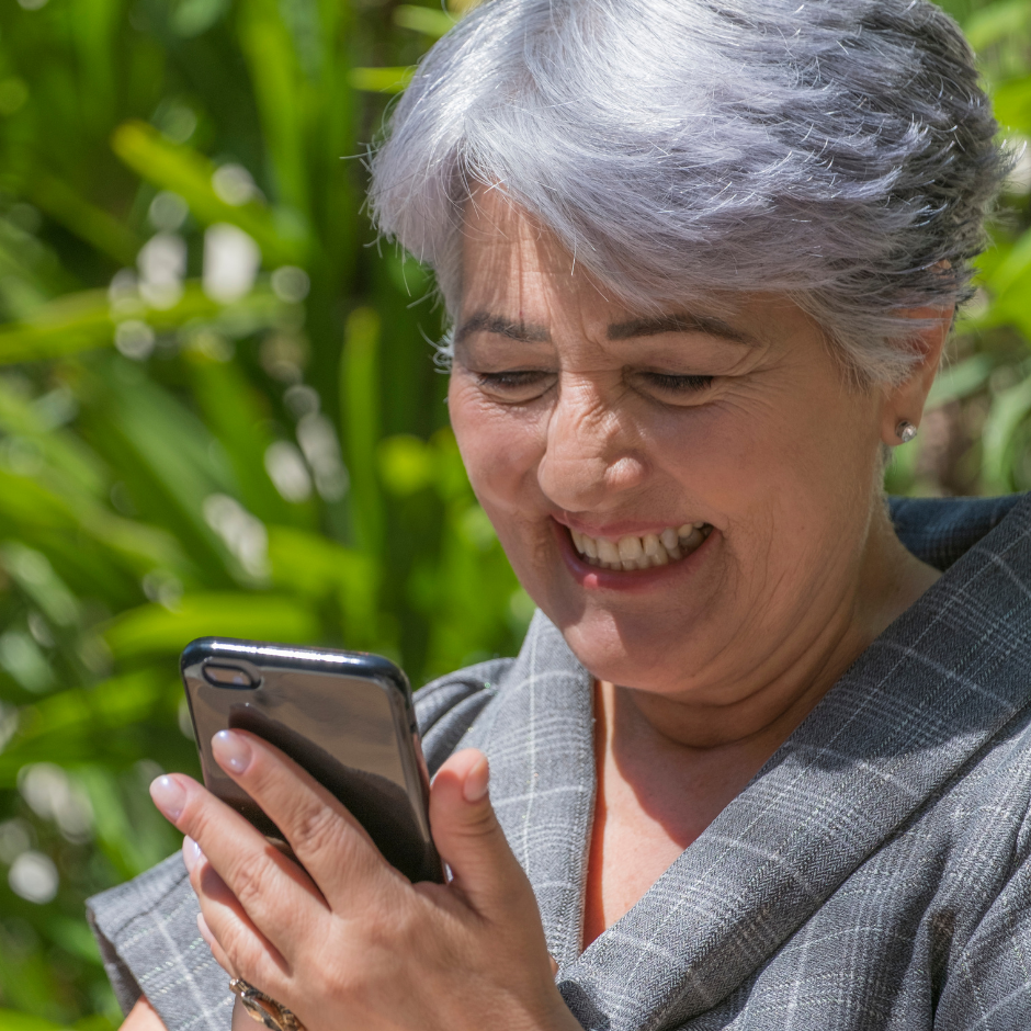 Foto de una mujer mayor de 60 años sonriendo y mirando la pantalla de su teléfono móvil, que sostiene en sus manos.