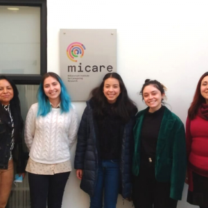 Foto de la visita de Itzel a las oficinas de MICARE. SE ven cinco mujeres jóvenes de pie sonriendo a la cámara. Detrás hay un letrero de MICARE.