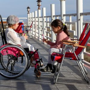 Foto que muestra a dos niñas jugando, una en su silla de ruedas y otra sentada en una silla de playa frente a ella.