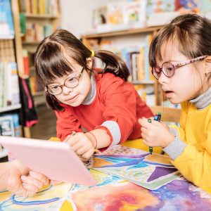 Foto de dos niñas con síndrome de down sentadas en una mesa en lo que parece ser una biblioteca, mirando una tablet. Una de ellas sostiene un crayón de color.