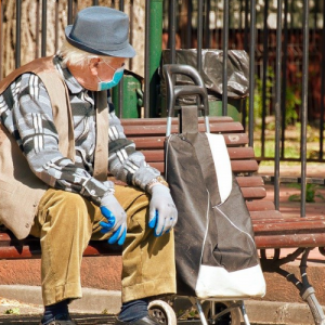 Foto de un hombre mayor con mascarilla y guantes sentado en una banca al sol, junto a un carro para hacer compras.