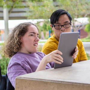 Foto de dos personas con discapacidad intelectual, una mujer y un hombre, mirando una tablet. Ambos están sentados en un parque.