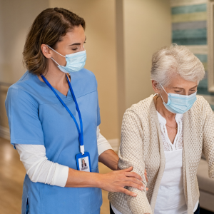 Foto de una mujer profesional de la salud con mascarilla, que ayuda a caminar a una mujer mayor también con mascarilla. Ambas están en un pasillo de un centro de salud.