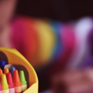 Foto de lápices de cera de colores. Detrás, borrosa, se ve una niña pintando con lápices.