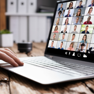 Foto de la mano de una persona junto a un computador portátil. En la pantalla se ven rostros de muchas personas en videollamada.